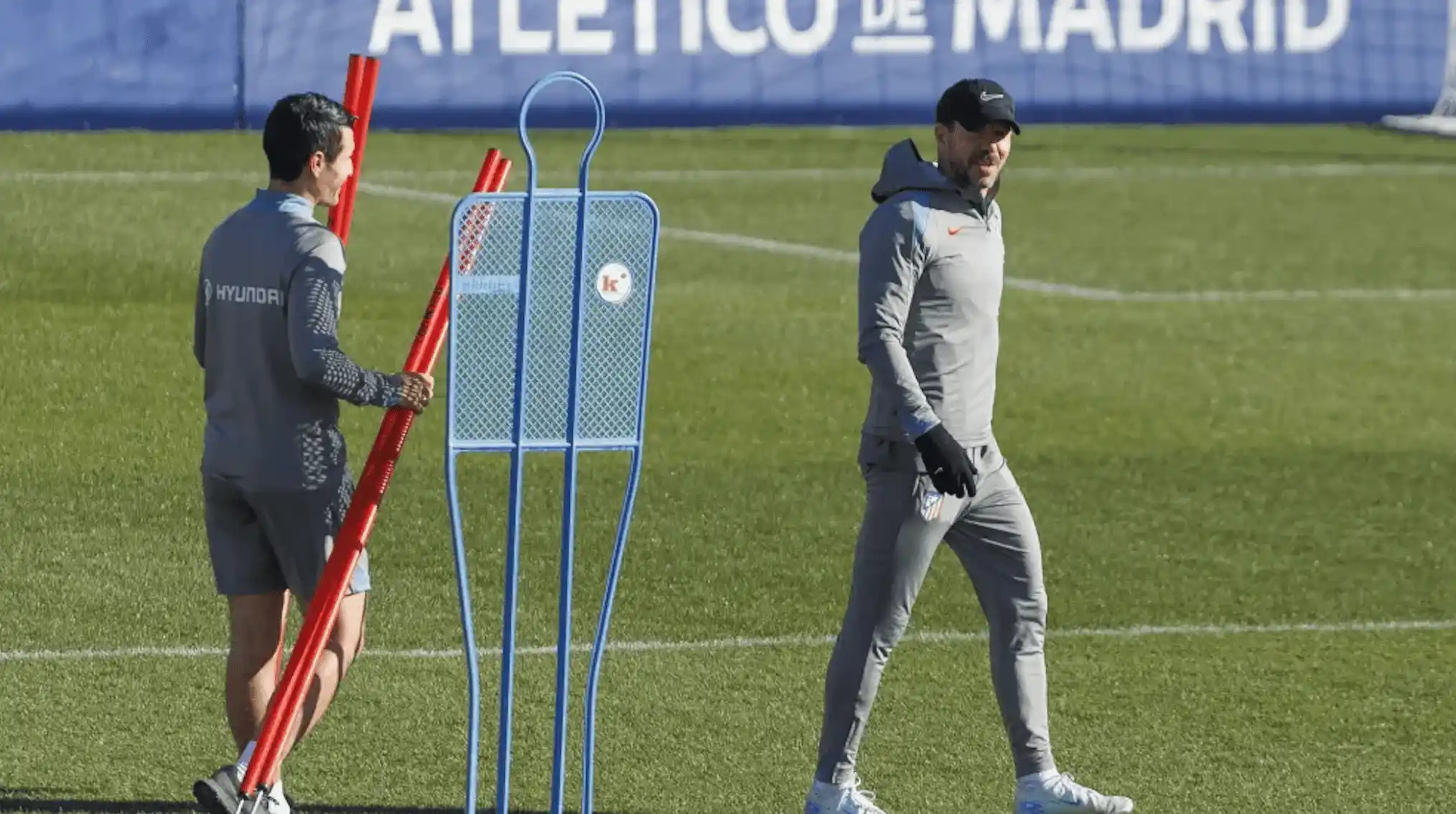 Entra&icirc;neurs de l'Atl&eacute;tico de Madrid sur un terrain d'entra&icirc;nement, l'un portant des jalons rouges et l'autre marchant en surv&ecirc;tement gris, &agrave; c&ocirc;t&eacute; d'un mannequin d'entra&icirc;nement en m&eacute;tal.